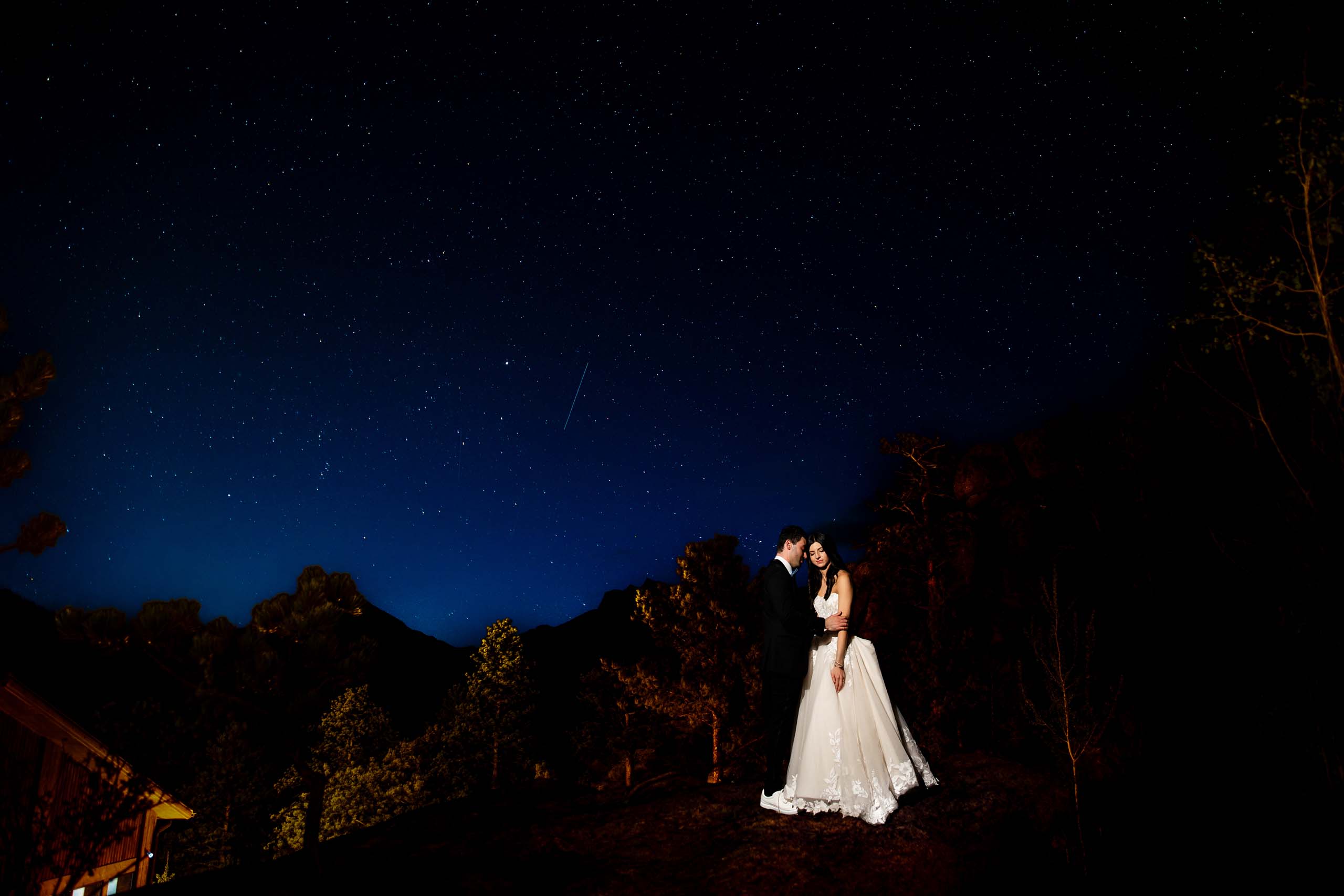Bride and groom in wedding attire embrace on a hill at night under a star filled sky with silhouettes of trees and mountains around them Colorado Wedding Photographer Colorado Wedding Photographer